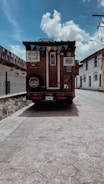 A rustic wooden mobile home is parked at the side of a quiet street with minimal traffic. The home has a triangular roof adorned with colorful pennant flags, and there are small windows on the back. The street is lined with traditional buildings and a stone wall, and the sky is partly cloudy, indicating a sunny day.