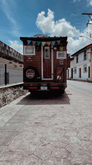 A rustic wooden mobile home is parked at the side of a quiet street with minimal traffic. The home has a triangular roof adorned with colorful pennant flags, and there are small windows on the back. The street is lined with traditional buildings and a stone wall, and the sky is partly cloudy, indicating a sunny day.