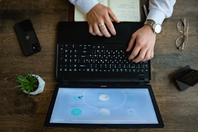 A person is typing on a black laptop placed on a wooden desk. Beside the laptop, there is a smartphone, a small potted plant, glasses, a notepad, and a business card holder. The screen displays circular graphics and text.