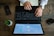 A person is typing on a black laptop placed on a wooden desk. Beside the laptop, there is a smartphone, a small potted plant, glasses, a notepad, and a business card holder. The screen displays circular graphics and text.