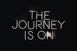 Neon lettering spells out 'The Journey is On' against a dark background, creating a bold and striking visual effect.