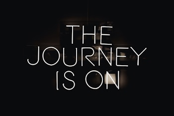 Neon lettering spells out 'The Journey is On' against a dark background, creating a bold and striking visual effect.
