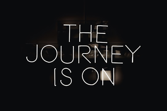 Neon lettering spells out 'The Journey is On' against a dark background, creating a bold and striking visual effect.