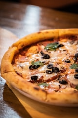 A close-up of a freshly baked pizza with a thick, golden-brown crust. The pizza is topped with melted cheese, black olives, slices of ham, and fresh basil leaves. It rests on a wooden surface, with warm tones creating a cozy and appetizing atmosphere.