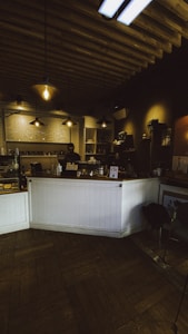 A cozy coffee shop interior with warm lighting and wooden ceilings. The counter is adorned with potted plants and a menu board is visible behind it. A person stands behind the counter, appearing to work. The space has a rustic feel with a combination of industrial lighting fixtures and wooden elements.