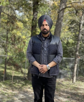 A man with a turban and beard stands in an outdoor setting, surrounded by trees and natural foliage. He is wearing a dark quilted vest over a plaid shirt, black pants, and a watch on his left wrist.