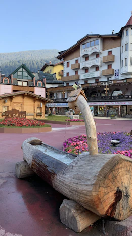 A rustic wooden fountain with water flowing from a spout is situated in a charming plaza. Surrounding the fountain are vibrant flowerbeds featuring purple and red blooms. A collection of buildings with Alpine architectural elements like balconies and pitched roofs encircle the plaza, under a clear blue sky with a hint of forested hills in the background.