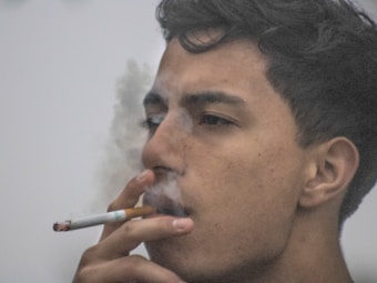 A person is seen in profile, inhaling on a cigarette with smoke curling around their face. The focus is on the facial expression and the act of smoking, set against a neutral background.