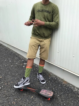A person stands against a white wall wearing an olive green sweatshirt with the word 'California' printed on it, khaki shorts, and vibrant green and black striped socks. The individual is also wearing black and white sneakers. At their feet is a skateboard with red wheels and patterned grip tape on a black and red deck.