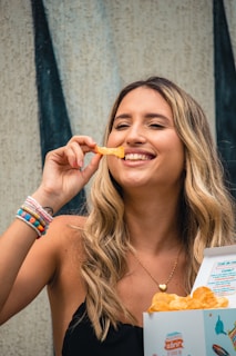 A woman with long blonde hair is smiling and holding a potato chip near her mouth. She is wearing a black top and several colorful bracelets on her wrist. In her other hand, she holds an open box of chips. The background features a textured wall with vertical black stripes.