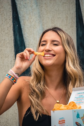 A woman with long blonde hair is smiling and holding a potato chip near her mouth. She is wearing a black top and several colorful bracelets on her wrist. In her other hand, she holds an open box of chips. The background features a textured wall with vertical black stripes.