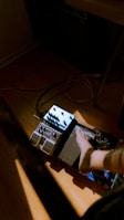A foot is resting on a guitar effects pedal board, which is placed on a wooden floor. Several pedals, including a volume pedal and multiple effects pedals with knobs and switches, are connected by wires. The scene is partially lit by natural sunlight, creating a warm and casual atmosphere.