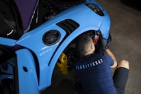 A person is working on a blue car, specifically the wheel area, with automotive tools. The individual is wearing a shirt with the word 'CLARITYCOAT' on the back. The car's front fender is visible, and the environment suggests a workshop setting.