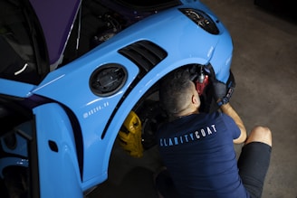 A person is working on a blue car, specifically the wheel area, with automotive tools. The individual is wearing a shirt with the word 'CLARITYCOAT' on the back. The car's front fender is visible, and the environment suggests a workshop setting.