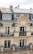A classic Parisian-style apartment building with ornate architectural details, wrought iron balconies adorned with potted plants and flowers, and beige stone facade. The top floor has a slate roof with dormer windows and red chimney stacks.