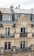 A classic Parisian-style apartment building with ornate architectural details, wrought iron balconies adorned with potted plants and flowers, and beige stone facade. The top floor has a slate roof with dormer windows and red chimney stacks.