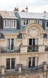 A classic Parisian-style apartment building with ornate architectural details, wrought iron balconies adorned with potted plants and flowers, and beige stone facade. The top floor has a slate roof with dormer windows and red chimney stacks.