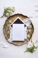 An elegant wedding invitation set placed on an ornate green and gold tray, surrounded by vintage postage stamps and soft white florals. Delicate white ribbons enhance the sophisticated arrangement, which is set against a light background.