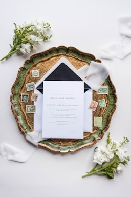 An elegant wedding invitation set placed on an ornate green and gold tray, surrounded by vintage postage stamps and soft white florals. Delicate white ribbons enhance the sophisticated arrangement, which is set against a light background.