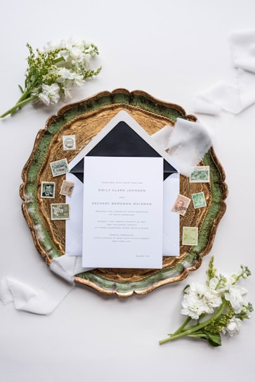 An elegant wedding invitation set placed on an ornate green and gold tray, surrounded by vintage postage stamps and soft white florals. Delicate white ribbons enhance the sophisticated arrangement, which is set against a light background.