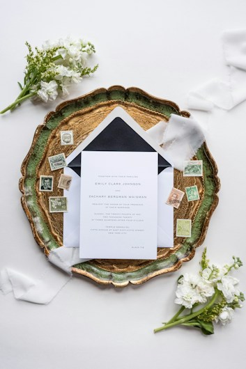 An elegant wedding invitation set placed on an ornate green and gold tray, surrounded by vintage postage stamps and soft white florals. Delicate white ribbons enhance the sophisticated arrangement, which is set against a light background.