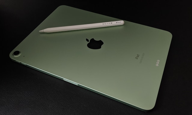 A green tablet lies on a dark surface with a white stylus placed diagonally across its back. The device has a black logo at the center and a camera in the top left corner.