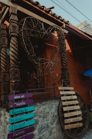 A rustic storefront features a large, heart-shaped ornament made of intricately woven branches, mounted on ornately carved wooden pillars. Below, several colorful signs list various drinks and food items in Spanish, such as Mezcal, Tequila, Micheladas, and Clericot. A stone wall supports the structure, and an old wooden wheel is repurposed for more signage.