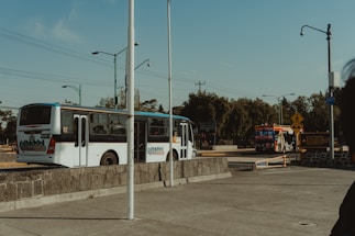 Two buses are on a street, one closer and one in the background. The closer bus is white with blue accents and has 'Ruta 4' written on it. The street is lined with various streetlights and utility poles. There are road signs, trees, and some buildings in the background. The sky is clear and blue.