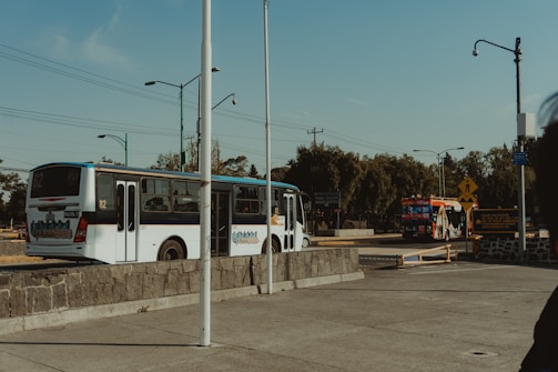 Two buses are on a street, one closer and one in the background. The closer bus is white with blue accents and has 'Ruta 4' written on it. The street is lined with various streetlights and utility poles. There are road signs, trees, and some buildings in the background. The sky is clear and blue.