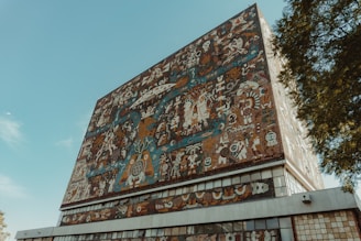 A large building facade adorned with a colorful, intricate mural featuring abstract and symbolic designs. Various figures, patterns, and shapes are interwoven, creating a dense visual tapestry. The mural displays rich detail with cultural and artistic elements.