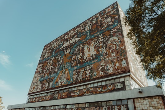 A large building facade adorned with a colorful, intricate mural featuring abstract and symbolic designs. Various figures, patterns, and shapes are interwoven, creating a dense visual tapestry. The mural displays rich detail with cultural and artistic elements.