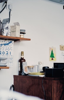 A kitchen corner with wooden shelves and a cabinet displaying various items including packets of food, a bottle, and a small calendar. A colorful drawing of a Christmas tree hangs on the wall, and a few plates are stacked neatly.