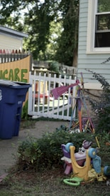 An outdoor scene featuring a corner of a building with a light blue siding and a window. Nearby, there's a colorful children's toy or playground equipment overturned on the grass. A bright pink object, possibly another toy, stands out among surrounding shrubs. A blue recycling bin is located near a white picket fence, with the word 'Care' partially visible on a colorful fence behind it. Mature trees and more greenery can be seen in the background, suggesting a backyard or garden setting.