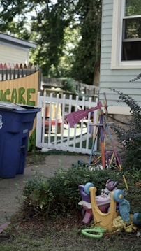 An outdoor scene featuring a corner of a building with a light blue siding and a window. Nearby, there's a colorful children's toy or playground equipment overturned on the grass. A bright pink object, possibly another toy, stands out among surrounding shrubs. A blue recycling bin is located near a white picket fence, with the word 'Care' partially visible on a colorful fence behind it. Mature trees and more greenery can be seen in the background, suggesting a backyard or garden setting.
