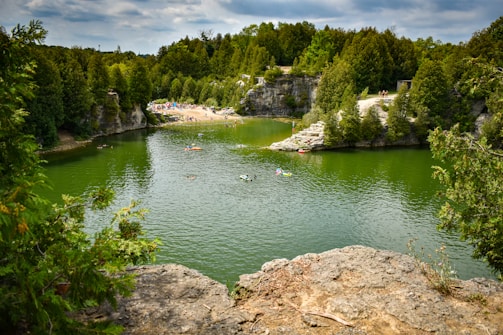 A lush, green landscape surrounds a tranquil body of water with several people enjoying leisure activities such as swimming and kayaking. The shoreline is lined with dense trees and rocky formations, while a sandy beach area can be seen with more people gathered.