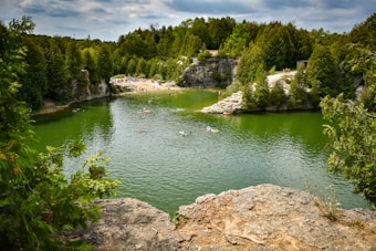 A lush, green landscape surrounds a tranquil body of water with several people enjoying leisure activities such as swimming and kayaking. The shoreline is lined with dense trees and rocky formations, while a sandy beach area can be seen with more people gathered.