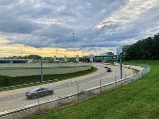A highway interchange with directional signs for Carmel and Indianapolis. Several cars travel on the road, which is surrounded by grassy areas and bordered by a wire fence. Office buildings are visible in the background, complemented by a cloudy sky with hints of a sunset.