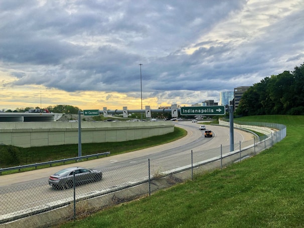 A highway interchange with directional signs for Carmel and Indianapolis. Several cars travel on the road, which is surrounded by grassy areas and bordered by a wire fence. Office buildings are visible in the background, complemented by a cloudy sky with hints of a sunset.
