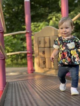 A young child with light hair, wearing a floral shirt and jeans, is joyfully walking on a playground structure. Vibrant pink columns and a number puzzle game are visible in the background, suggesting a lively play environment surrounded by lush greenery.