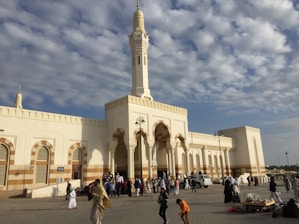 A large mosque with a tall minaret stands against a partly cloudy sky. The building is characterized by its white and beige exterior with ornate archways and geometric patterns. Several people are gathered in the courtyard, some walking, and others engaged in conversation. The atmosphere is lively, suggesting a congregational setting.