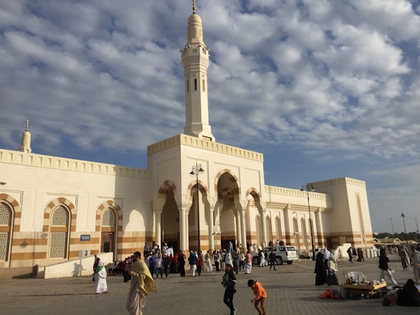 A large mosque with a tall minaret stands against a partly cloudy sky. The building is characterized by its white and beige exterior with ornate archways and geometric patterns. Several people are gathered in the courtyard, some walking, and others engaged in conversation. The atmosphere is lively, suggesting a congregational setting.