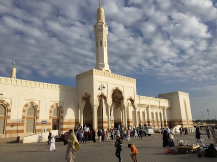 A large mosque with a tall minaret stands against a partly cloudy sky. The building is characterized by its white and beige exterior with ornate archways and geometric patterns. Several people are gathered in the courtyard, some walking, and others engaged in conversation. The atmosphere is lively, suggesting a congregational setting.