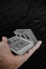 A hand holds a deck of playing cards with intricate back designs and a visible five of clubs. The background is a dark, textured surface, contrasting with the lighter tones of the cards and hand.