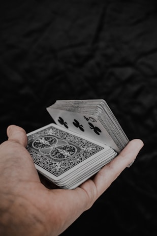 A hand holds a deck of playing cards with intricate back designs and a visible five of clubs. The background is a dark, textured surface, contrasting with the lighter tones of the cards and hand.