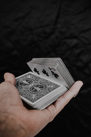 A hand holds a deck of playing cards with intricate back designs and a visible five of clubs. The background is a dark, textured surface, contrasting with the lighter tones of the cards and hand.