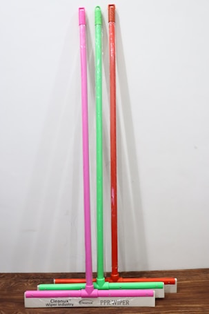 Four squeegees with long handles in pink, green, and red colors are standing upright against a white background. The base of each squeegee is white with text printed on it. They are arranged in a line on a wooden surface.