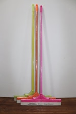 Three squeegees are lined up against a plain wall on a wooden surface. Each has a brightly colored handle in neon shades of yellow, orange, and pink. The squeegee blades and handles are wrapped in clear plastic packaging. The base of each squeegee has the branding 'Cleanux Wiper Industry' and 'King Master' printed on it.