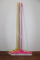 Three squeegees are lined up against a plain wall on a wooden surface. Each has a brightly colored handle in neon shades of yellow, orange, and pink. The squeegee blades and handles are wrapped in clear plastic packaging. The base of each squeegee has the branding 'Cleanux Wiper Industry' and 'King Master' printed on it.