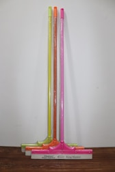 Three squeegees are lined up against a plain wall on a wooden surface. Each has a brightly colored handle in neon shades of yellow, orange, and pink. The squeegee blades and handles are wrapped in clear plastic packaging. The base of each squeegee has the branding 'Cleanux Wiper Industry' and 'King Master' printed on it.