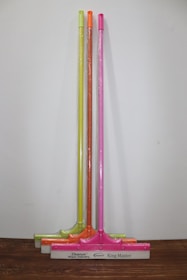 Three squeegees are lined up against a plain wall on a wooden surface. Each has a brightly colored handle in neon shades of yellow, orange, and pink. The squeegee blades and handles are wrapped in clear plastic packaging. The base of each squeegee has the branding 'Cleanux Wiper Industry' and 'King Master' printed on it.